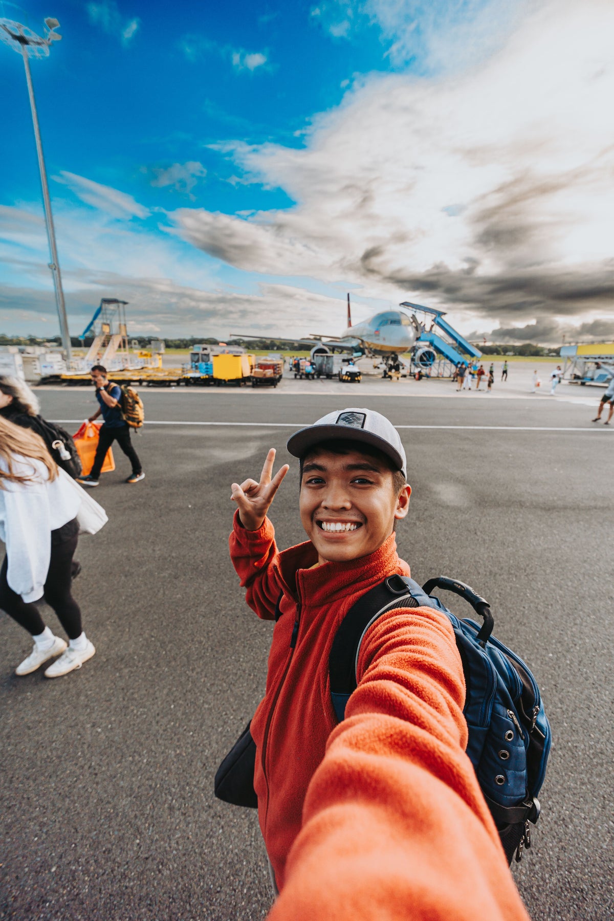 A man taking a selfie on an airport tarmac, wearing a bright orange fleece jacket, a gray baseball cap, and a blue backpack, with airplanes and a blue sky in the background.